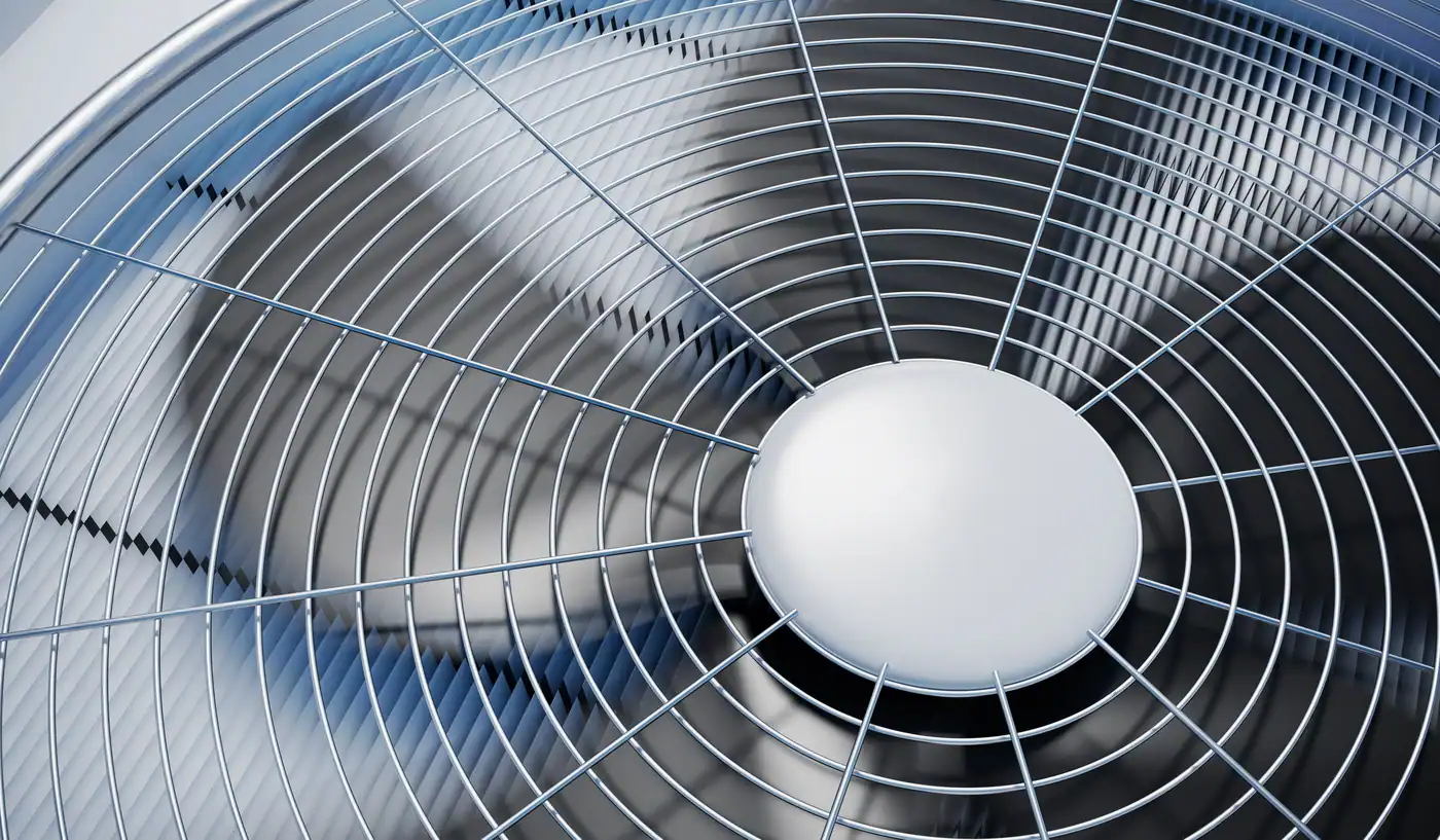 Close-up image of a metal fan inside HVAC unit with curved blades and protective grille. The fan is large, occupying most of the frame, and appears to be in a sunny outdoor setting, as indicated by the reflections on the blades.