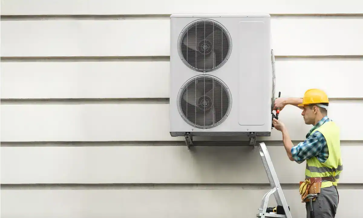 A construction worker wearing a yellow hard hat and safety gear stands on a ladder and uses tools to provide HVAC service, installing or repairing a large, mounted air conditioning unit on the outside of a building.