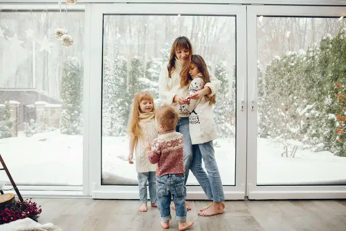 A woman and three children stand barefoot in a cozy room with large windows, showing a snowy outdoor scene. The woman and two girls are smiling and embracing, while a toddler faces them with curiosity after the electrician finished his work ensuring their comfort. They are all dressed warmly in sweaters and jeans.