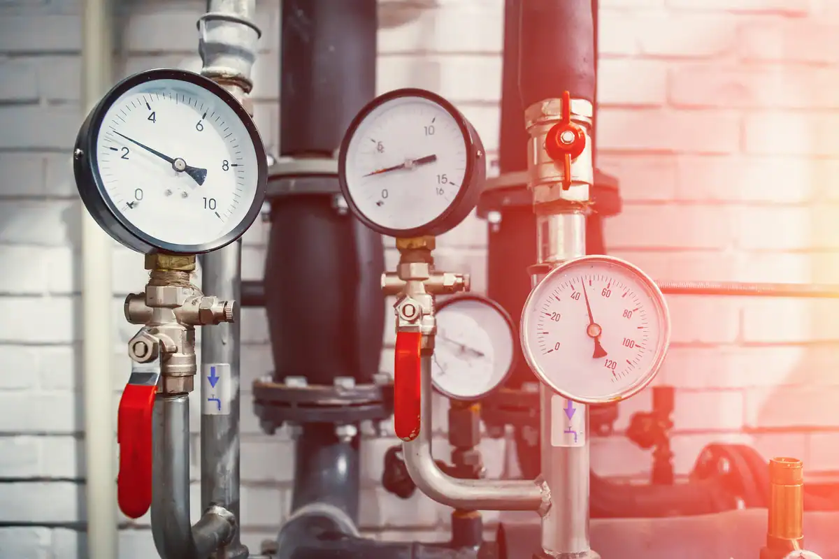 Three industrial pressure gauges with red handles are mounted on pipes against a brick wall background. The gauges display various pressure readings, suggesting a mechanical or furnace repair setting. Sunlight is seen shining from the right side of the image.