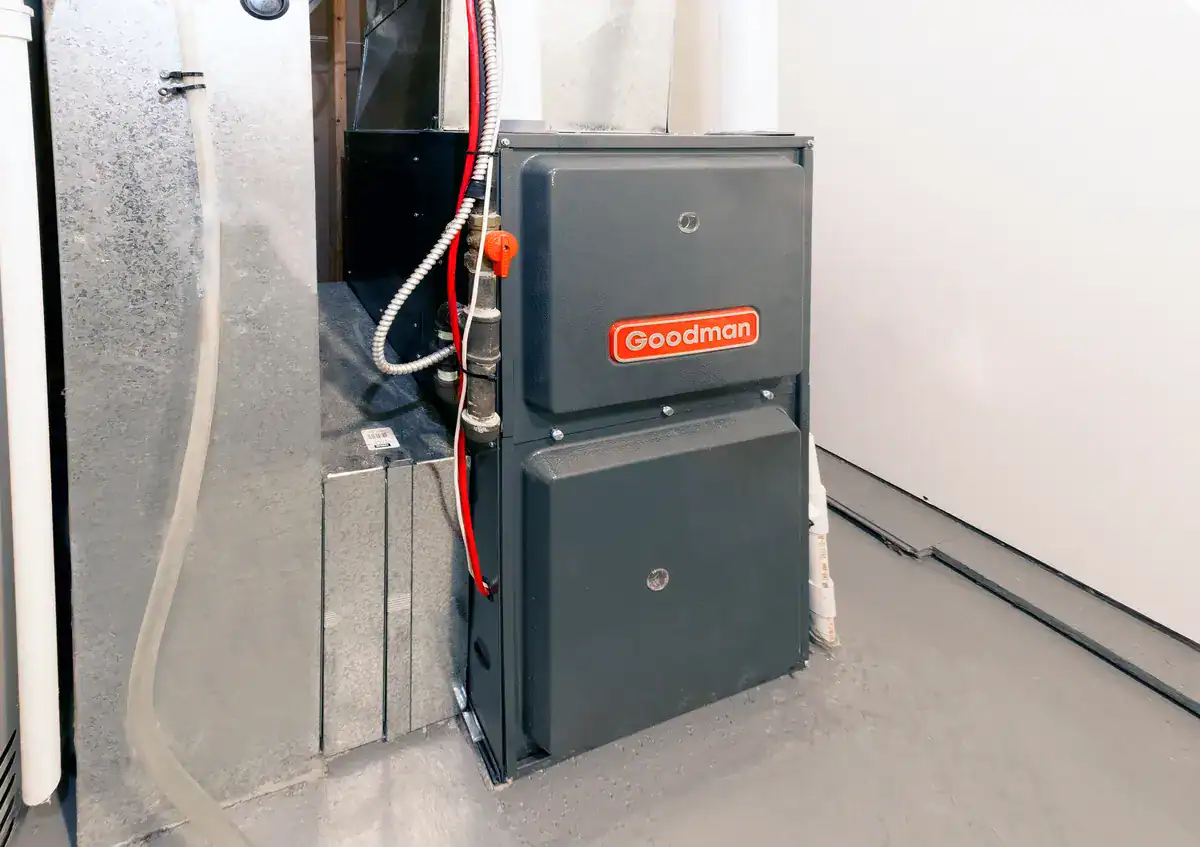 A gray Goodman furnace unit is installed on a cement floor in a utility room. It has metal ductwork connected at the top and electrical wiring attached on the side. The room has white walls and a neat appearance.