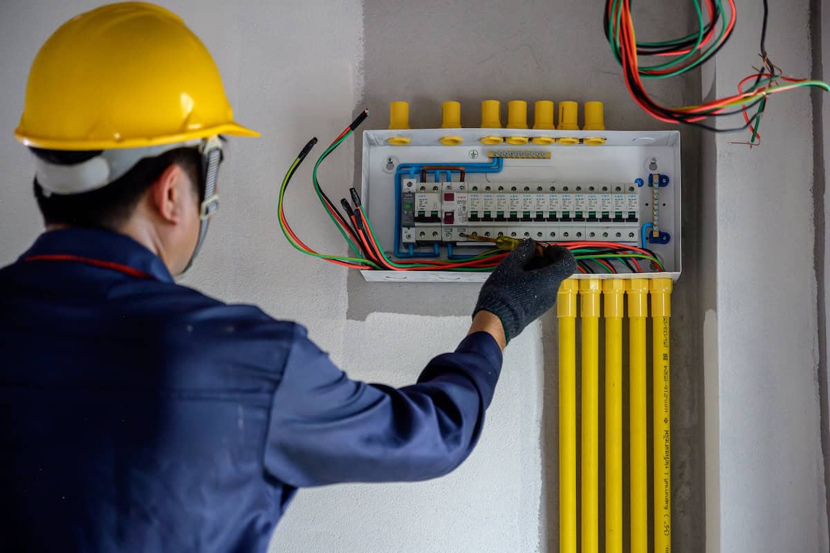 An electrician near me in Bellevue, wearing a yellow hard hat and dark blue uniform, inspects an electrical panel on the wall. Colorful wires extend from the panel, accompanied by several yellow conduit pipes running vertically below.