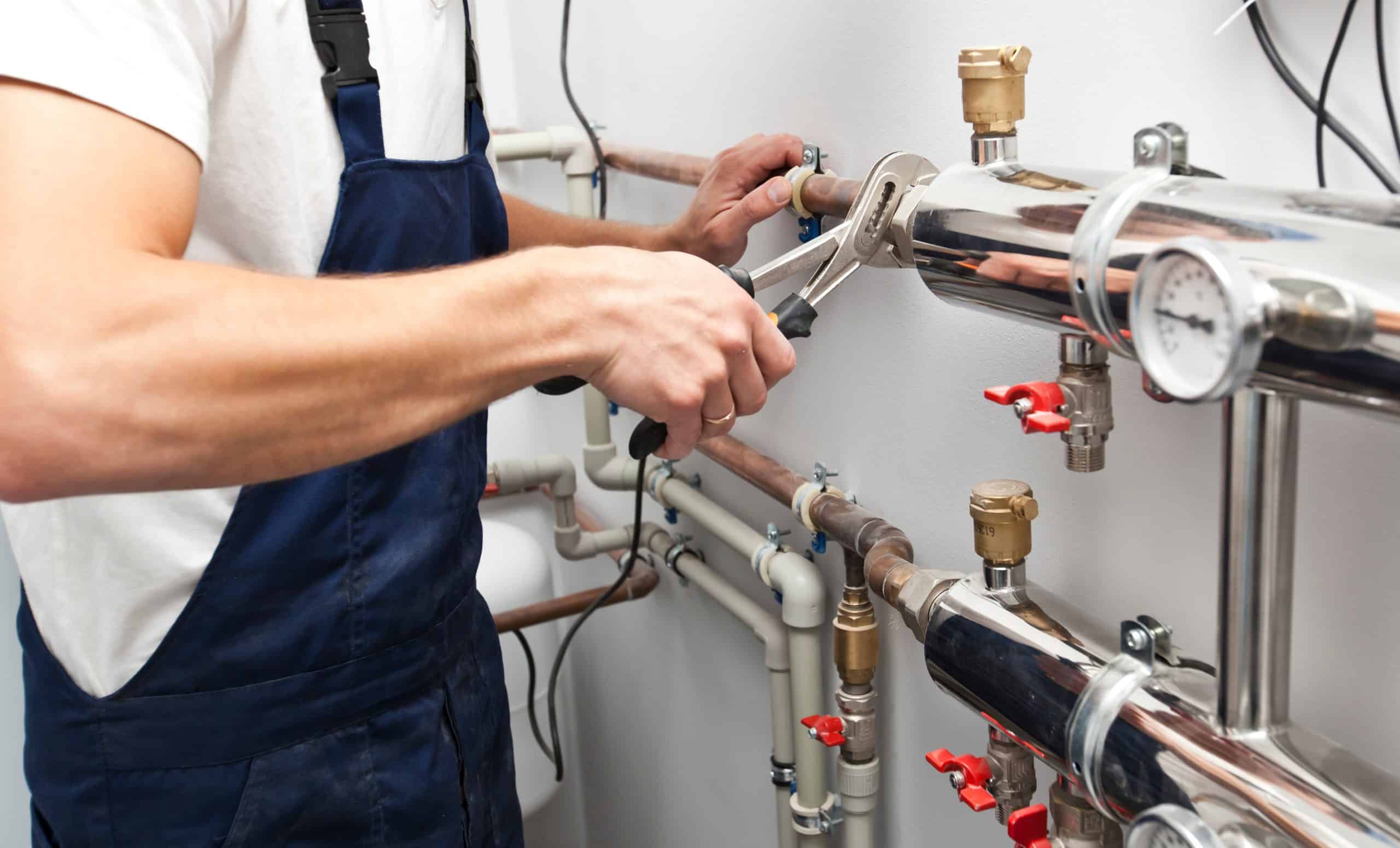 A local plumber in Appleton, WI, clad in blue overalls, expertly uses pliers on a heating system. The setup features a metallic pipe with valves and meters neatly installed on a pristine white wall.