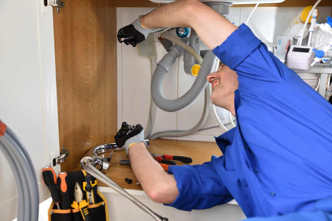 A person in a blue shirt is lying under a kitchen sink, expertly fixing plumbing for drain cleaning near me. They are using tools with a toolbox nearby, surrounded by various pipes, hoses, and cleaning supplies in the background, ensuring everything flows smoothly in South St. Paul MN.