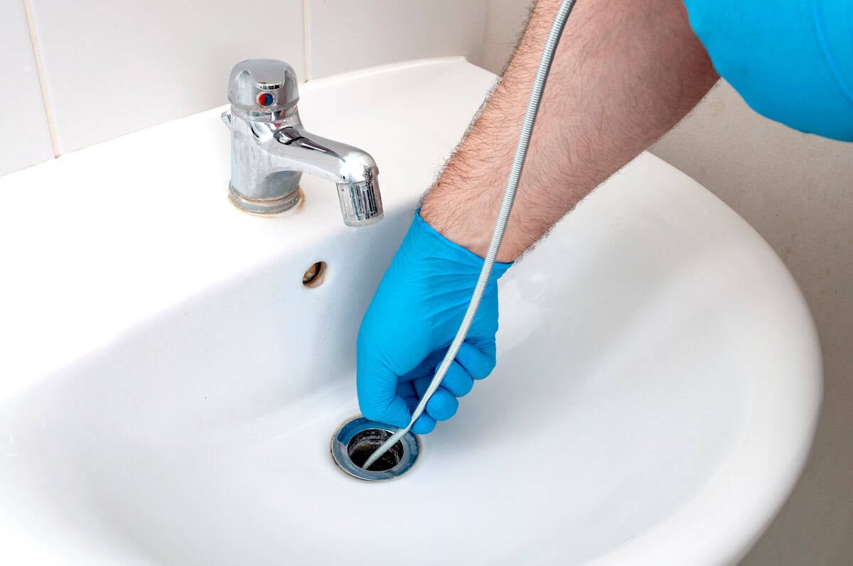 A person wearing blue gloves is using a plumbing snake to unclog a white sink. The snake is being inserted into the drain, and there is a metal faucet visible on the left. The setting is a bathroom with a tiled wall.
