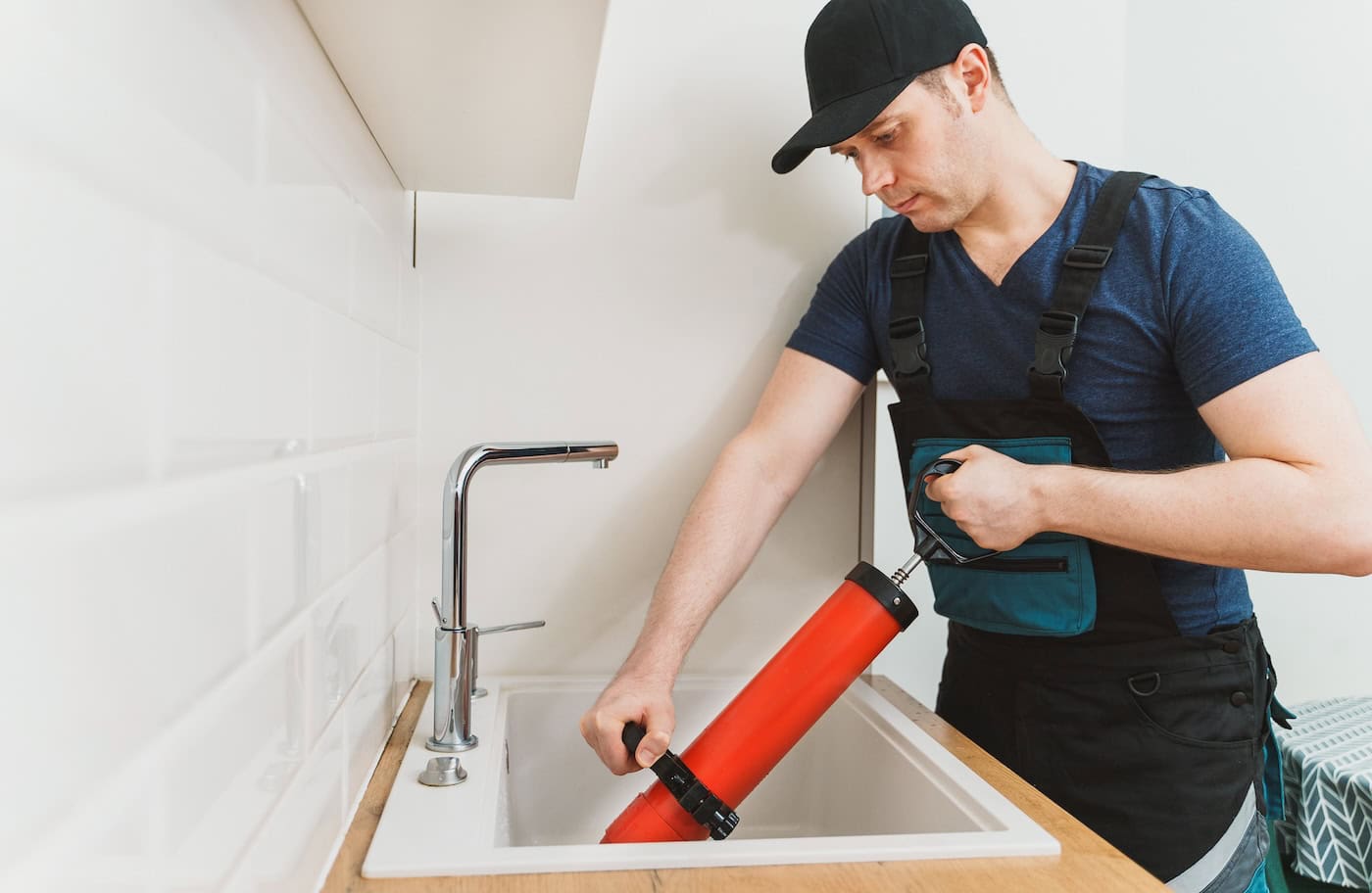 A plumber in a blue shirt and black cap uses a red plunger to unclog a kitchen sink. The setting is a clean, modern kitchen with white tiles and cabinetry.