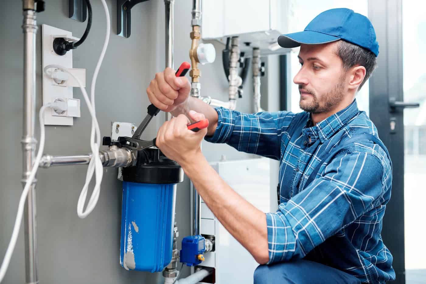 A local plumber from Menasha, WI, in a blue cap and striped shirt, is working on a water filtration system. He skillfully adjusts a blue filter with wrenches, surrounded by pipes in the utility room, while electrical outlets and various machinery hum quietly in the background.