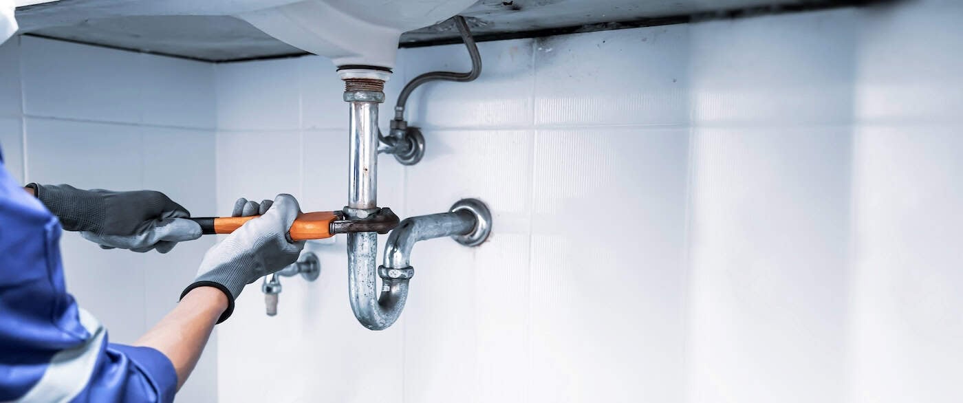 A plumber wearing gloves uses a wrench to tighten or loosen a pipe under a white sink, focusing on efficient drainage. The exposed plumbing, with metal pipes and fittings against the tiled wall backdrop, suggests he specializes in drain cleaning near Mendota Heights, MN.