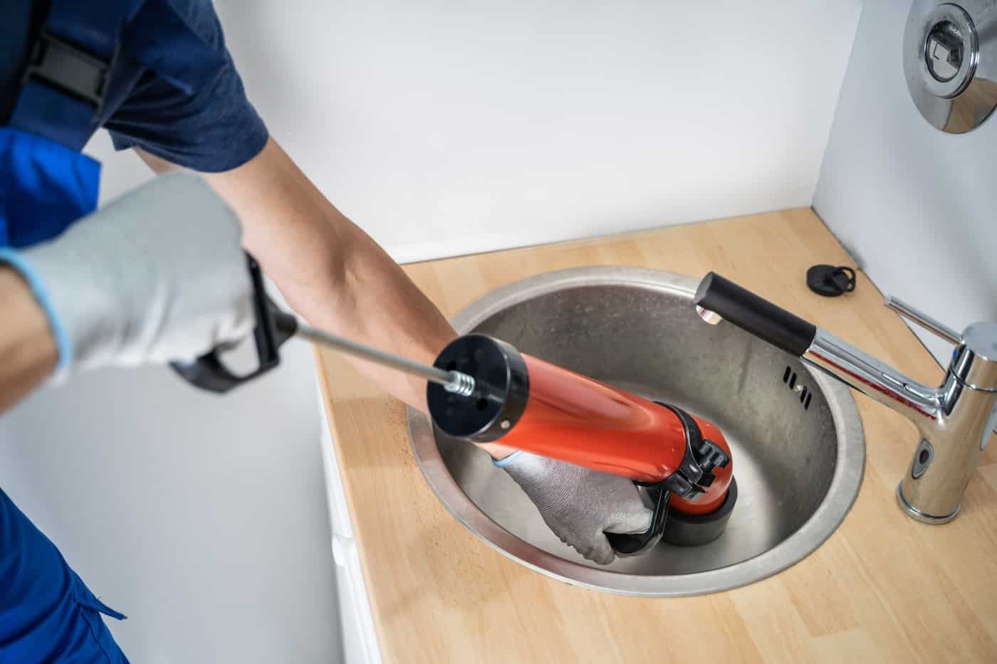 A plumber wearing gloves uses an orange plunger or drain tool to unclog a kitchen sink faucet, expertly handling the drain cleaning task. The wooden countertop and stainless steel sink are visible, evoking a typical home repair setting in Lakeville, MN.