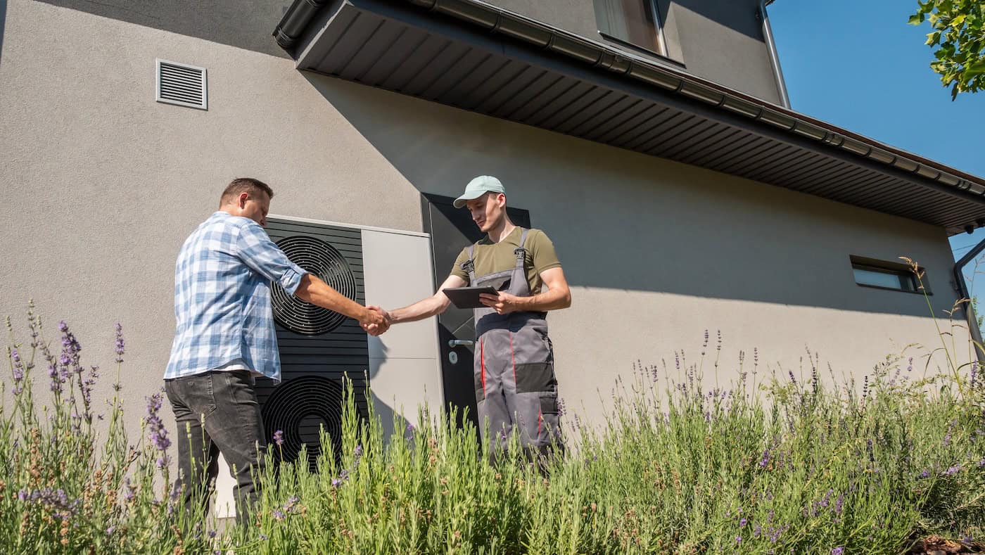 A Homeowner And A Technician Shake Hands In Front Of A House Near An External Heat Pump Unit.