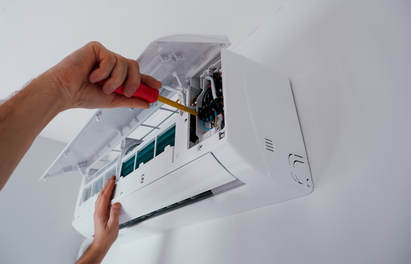 A person uses a screwdriver to repair or maintain the internal components of a wall-mounted air conditioner unit with its front panel open.