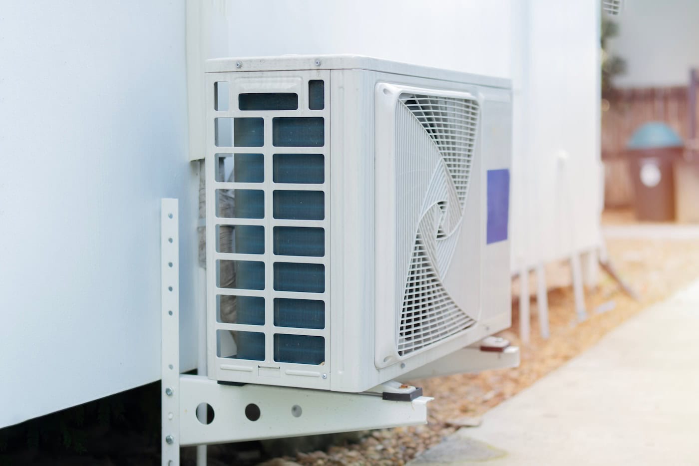Close-up of an outdoor hvac unit in Little Falls, MN, mounted on a metal bracket attached to the building’s exterior wall, with another unit and a pathway softly blurred in the background.