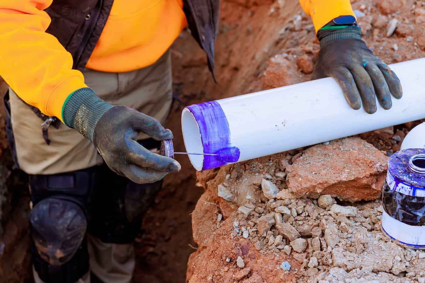 Worker Applies Purple Primer To Pvc Pipe In Construction Site During Daylight Hours In A Trench