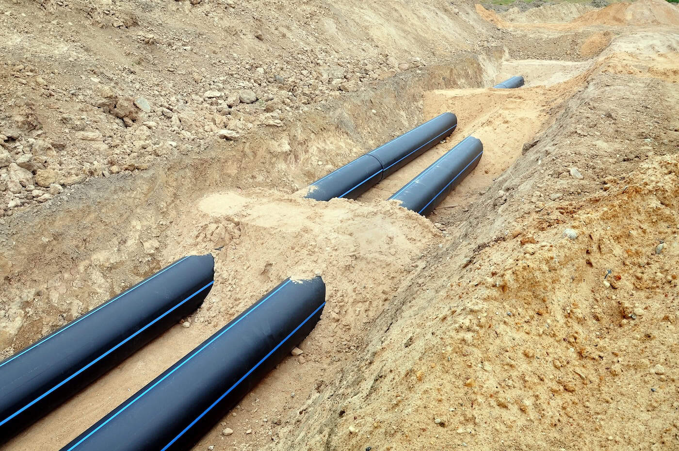 The Process Of Laying Of Engineering And Heating Systems. Two Black Plastic Pipes Are In A Trench Of Sand In Perspective.