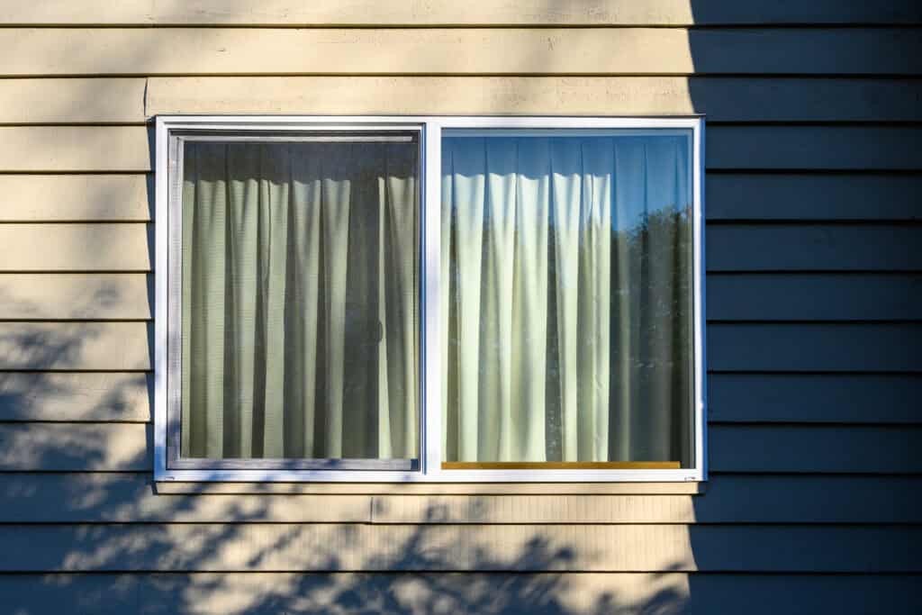 A sunlit, beige house wall with horizontal siding features a closed window. The window has glass panes and light green curtains, with shadows from nearby trees cast on the wall and window.