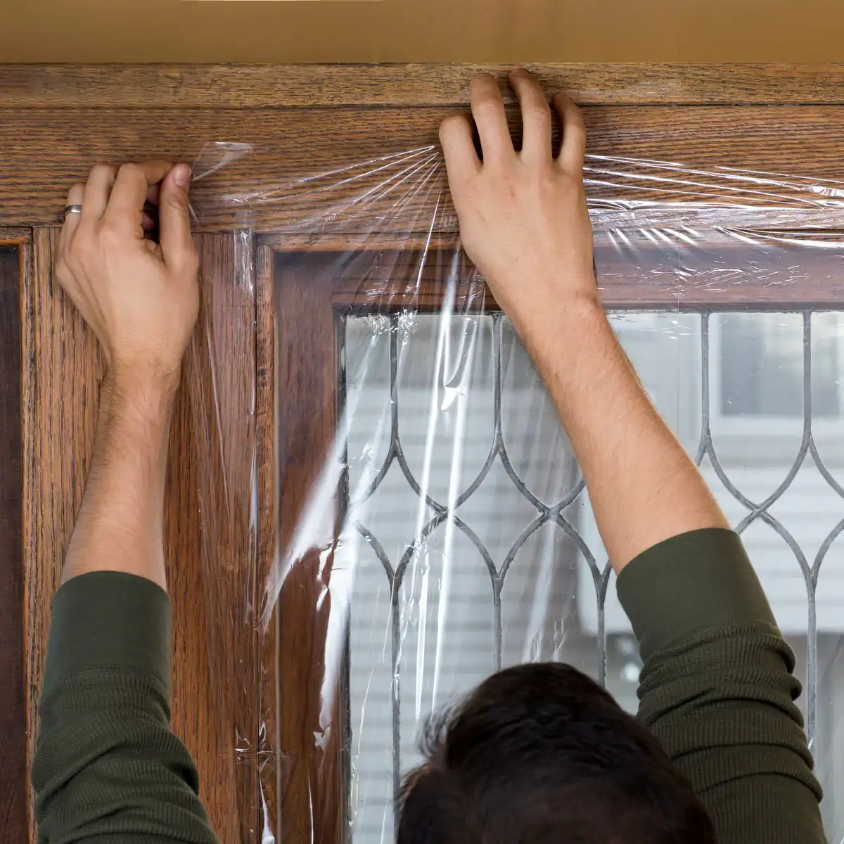 A person in a green long-sleeve shirt applies clear plastic film to a wooden framed window with decorative glass.
